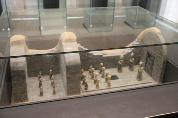 Ein Museumsdisplaycase voller verschiedener Artefakte, einschließlich Skulpturen und Glasboxen, vor einer Wand.