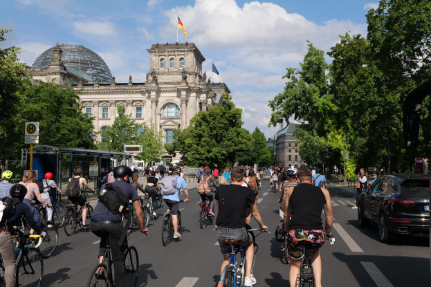 Eine Gruppe von Menschen, die auf Fahrrädern eine von Bäumen gesäumte Straße in Berlin, Deutschland, entlangfährt, mit einer Bushaltestelle auf der rechten Seite und einer Flagge, die auf einem nahegelegenen Gebäude weht.