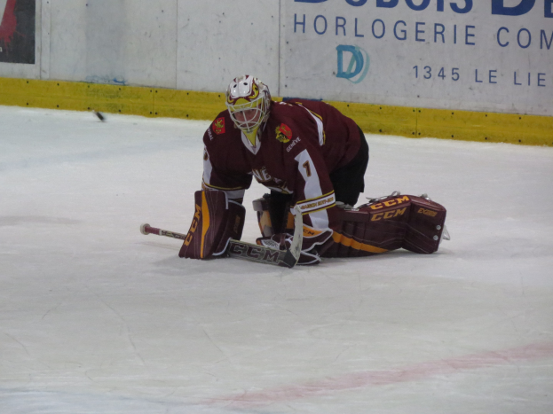 Eishockeyspieler in rot-gelber Uniform, der einen Schuss auf dem Eis abwehrt, mit einer Wand und Text im Hintergrund.