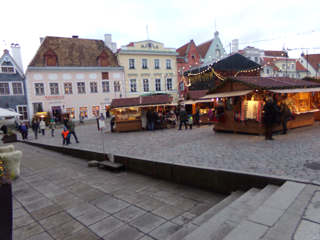 Ein lebendiger Weihnachtsmarkt in Tallinn, Estland, mit Menschen um beleuchtete und geschmückte Stände, Gebäuden mit Fenstern im Hintergrund, Treppen, die zum Markt führen, und Töpfen mit Pflanzen, die herumstehen.
