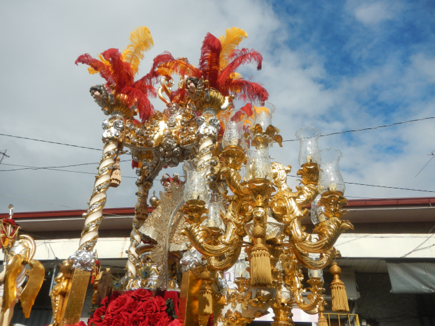 Ein großes goldenes und rotes Festwagen, geschmückt mit Blumen und anderen Dekorationsgegenständen, in einem Karnevalsumzug, mit einem Gebäude, Strommasten mit Drähten und einem bewölkten Himmel im Hintergrund.