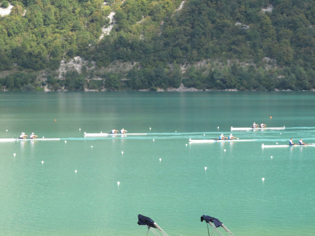 Gruppe von Menschen bei einem Ruderwettbewerb auf einem Gewässer mit Bäumen und einem Hügel im Hintergrund, Kleidung über Stühle am Boden drapiert.