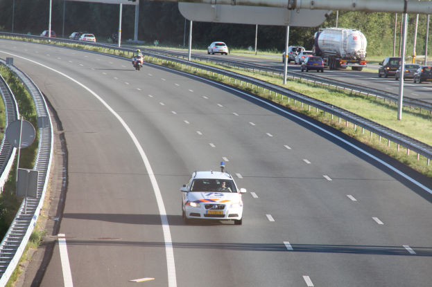 Ein Auto fährt auf einer Autobahn mit einem Polizeiwagen am Rande, anderen Fahrzeugen, Geländern, Polen, Platten, Brettern und Bäumen im Hintergrund.