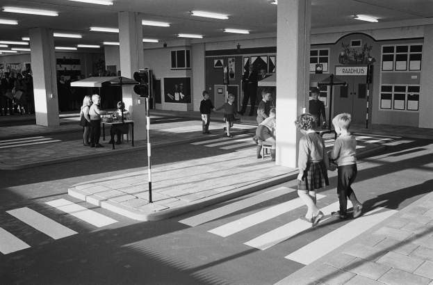 Schwarzes und weißes Foto von Kindern, die eine Straße an einer Ampel überqueren, mit Fußgängern in der Nähe, einem Verkehrssignalmast im Vordergrund und strukturellen Säulen und Leuchten im Hintergrund.