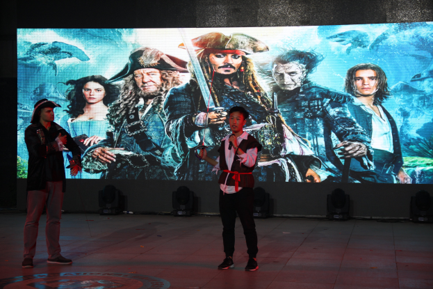 Zwei Personen stehen auf einer Bühne vor einem großen Bildschirm, eine hält ein Mikrofon und die andere ein Objekt, mit Scheinwerfern im Fokus und einem Pirates of the Caribbean-Plakat im Hintergrund.