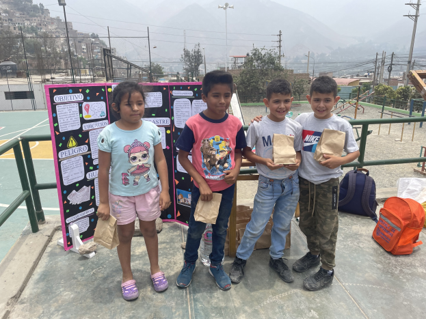 Gruppe von Kindern auf einem Basketballfeld mit Papierbeuteln in den Händen, mit Beuteln und Gegenständen rechts und einer Tafel mit Text links, vor einem Hintergrund aus Zäunen, Pfosten, Straßenlaternen, Bäumen, Gebäuden, Hügeln und Himmel.
