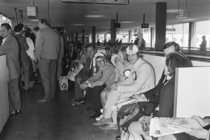 Eine Gruppe von Menschen verschiedenen Alters, einige stehend, einige sitzend, in einem Flughafenwartebereich mit Glasfenstern auf der rechten Seite, in Schwarz-Weiß.