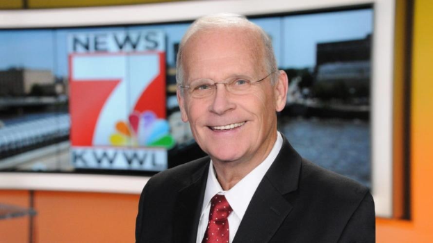 Ein Mann im Anzug und Krawatte, der eine Brille trägt, steht lächelnd vor einem Fernseher mit leicht unscharfem Hintergrund.