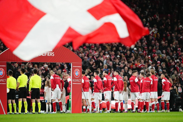 Eine Gruppe von Menschen auf einem Fußballfeld mit einer roten und weißen Fahne im Vordergrund, einem Bogen mit der Aufschrift "Bayern Munich vs Bayern Munich Betting Tips & Preview" im Hintergrund und einer großen Menge im Stadion.