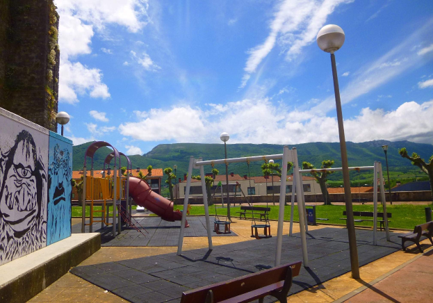 Spielplatz mit Schaukeln, Rutsche, Bänken, Masten, Lichtern, einer bemalten Wand, Gras, Bäumen, Gebäuden, Hühlen und einem bewölktem Himmel im Hintergrund.