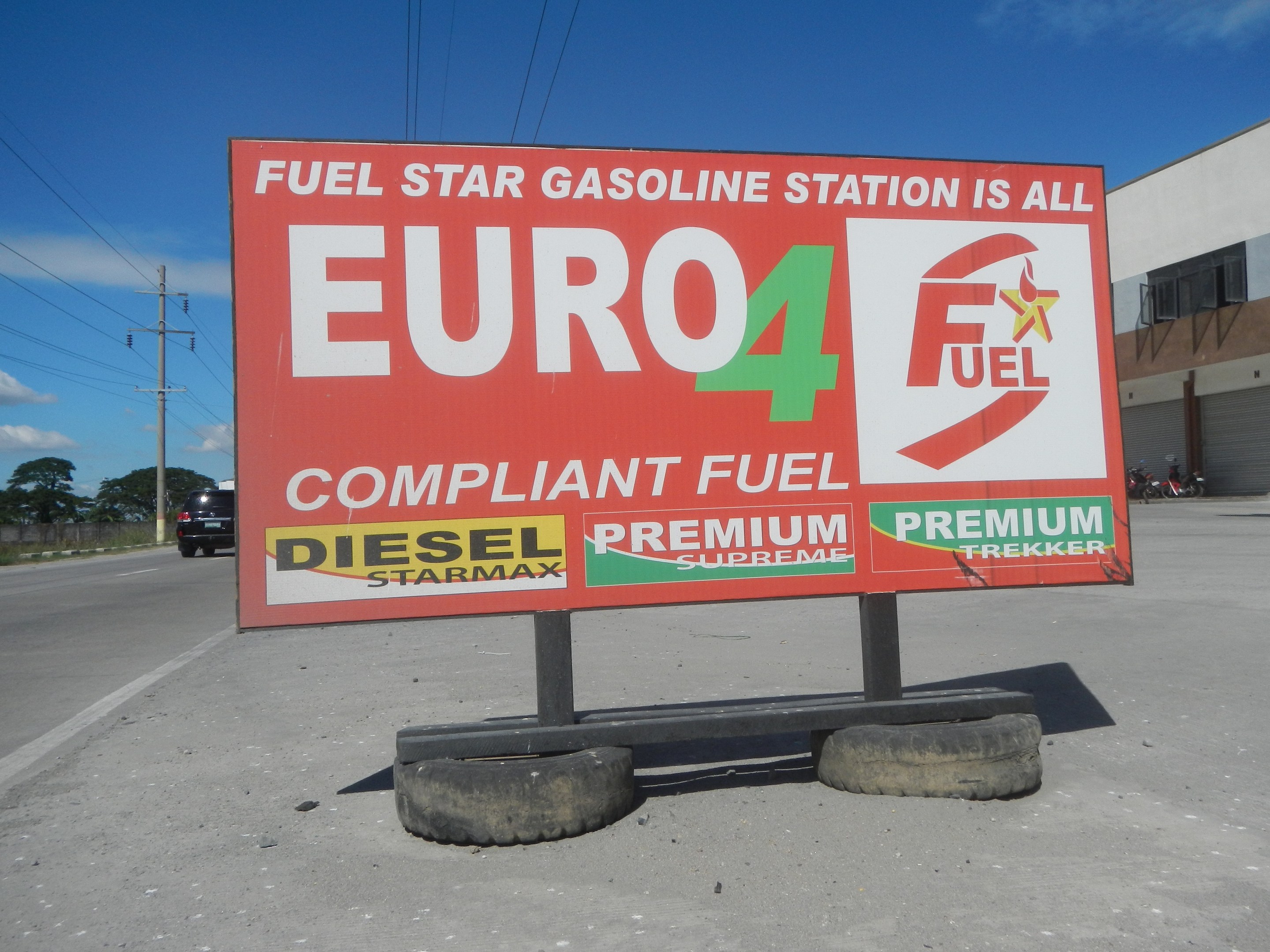 Eine Tankstelle mit einem "Euro4 Fuel"-Schild im Vordergrund, Fahrzeuge auf der Straße, Strommäste mit Drähten, Bäume, ein Gebäude auf der rechten Seite und ein sichtbarer Himmel.