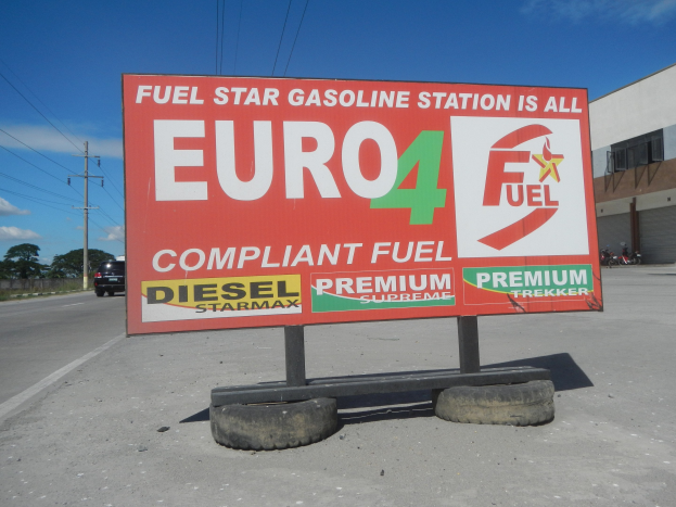 Eine Tankstelle mit einem "Euro4 Fuel"-Schild im Vordergrund, Fahrzeuge auf der Straße, Strommäste mit Drähten, Bäume, ein Gebäude auf der rechten Seite und ein sichtbarer Himmel.