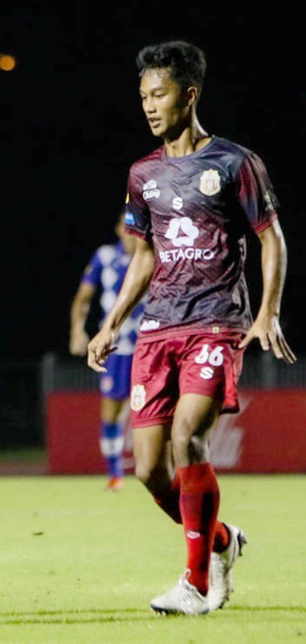 Ein professioneller Fußballspieler in einem rotbraunen T-Shirt und roten Shorts, der einen Ball auf einem Feld kickt, mit einer anderen Person im Hintergrund und einer Tafel mit Schrift.