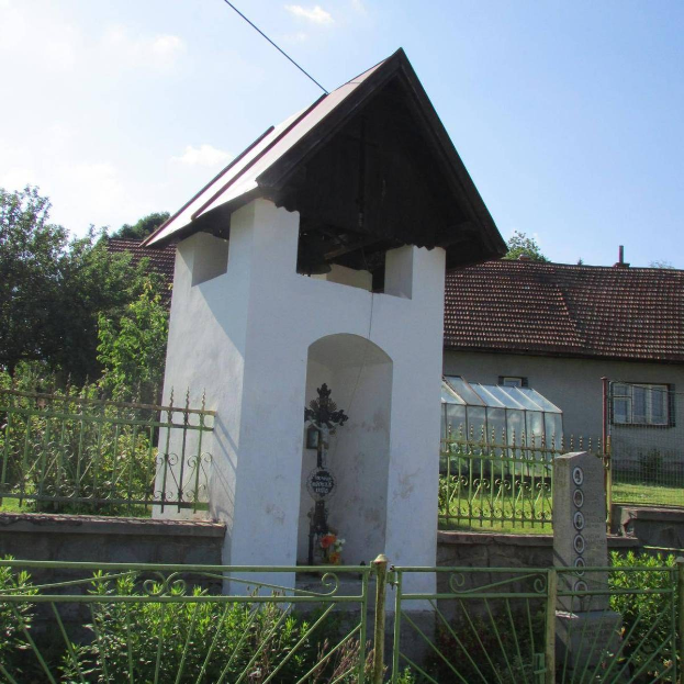 Kleine weiße Kirche mit einem Kreuz darauf, umgeben von einem Metallzaun, Pflanzen und einem Weg, vor Häusern und einem bewölkten Himmel.