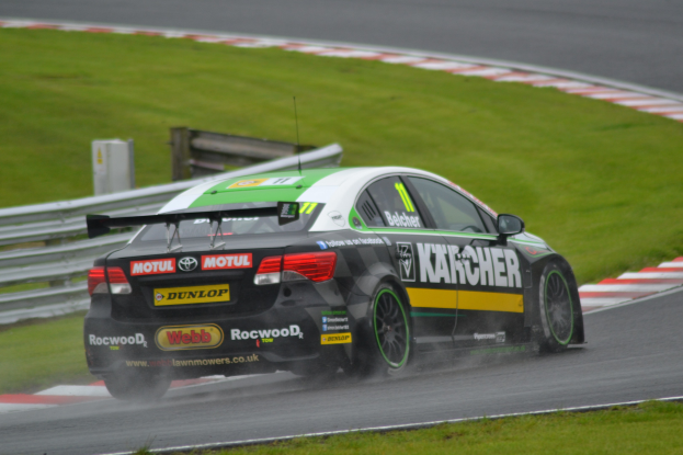 Ein Auto mit Text auf der Seite, das auf einer regennassen Rennstrecke fährt, die von Gras gesäumt ist, mit einem Geländer und einem hölzernen Gebäude im Hintergrund.