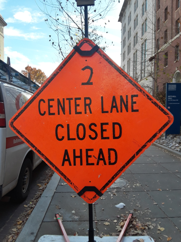 Orangefarbenes Schild mit der Aufschrift "2 Center Lane Closed Ahead" an der Seite einer Straße mit Fahrzeugen, Pfosten, Planken, Bäumen, Gebäuden und einem bewölkten Himmel im Hintergrund.