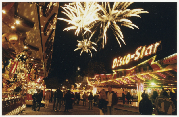A crowd of people stands in front of a brightly lit carnival ride at night, with buildings, trees, and fireworks in the background.