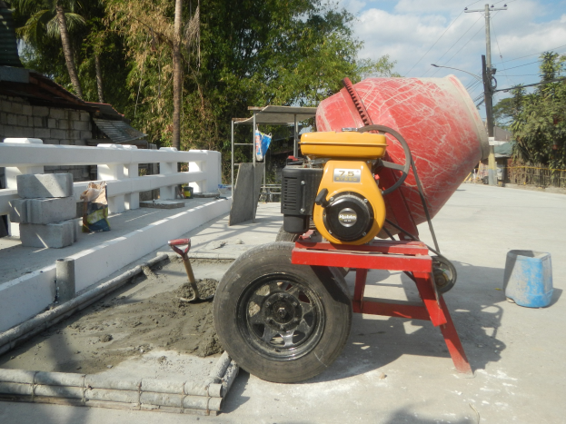 Betonmischer auf einem Zementlaster umgeben von Baustoffen, einem Haus, Bäumen, Strommasten und einem bewölkten Himmel.