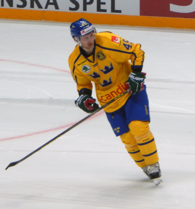 Eishockeyspieler in Bewegung auf dem Eis, mit Helm, Handschuhen und Schlittschuhen, einen Hockeystock haltend, mit einem Hintergrundboard mit Text.