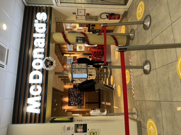 Innenseite eines McDonald's-Restaurants mit einem sichtbaren 'McDonald's'-Schild auf der linken Seite, roten Metallstangen mit Bändern auf der rechten Seite und einigen Menschen in der Nähe des Eingangs mit Hintergrundgegenständen.