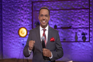 Stephen Colbert, der einen Anzug, eine Krawatte und eine Brille trägt, steht auf einer Bühne und hält ein Mikrofon in der Hand, während hinter ihm eine Wand und verschiedene Gegenstände zu sehen sind.