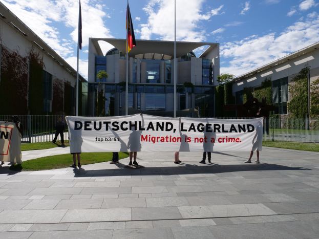 Eine Gruppe von Menschen hält ein Banner mit der Aufschrift "Deutschland, Lagerland Migration ist kein Verbrechen" vor einem eingezäunten Gebäude mit Bäumen und Flaggen.