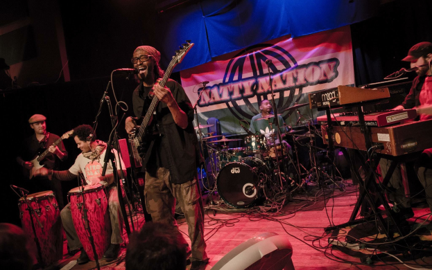 Eine Live-Band performt auf der Bühne mit Mikrofonen und Ständern, während das Publikum unten zuschaut; im Hintergrund ist ein Banner zu sehen.