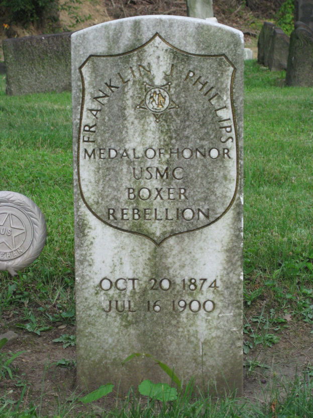 Ein Grabstein mit der Inschrift "Franklin J. Phillips Medal of Honor USMC Boxer Rebellion" umgeben von Gras und Bäumen.