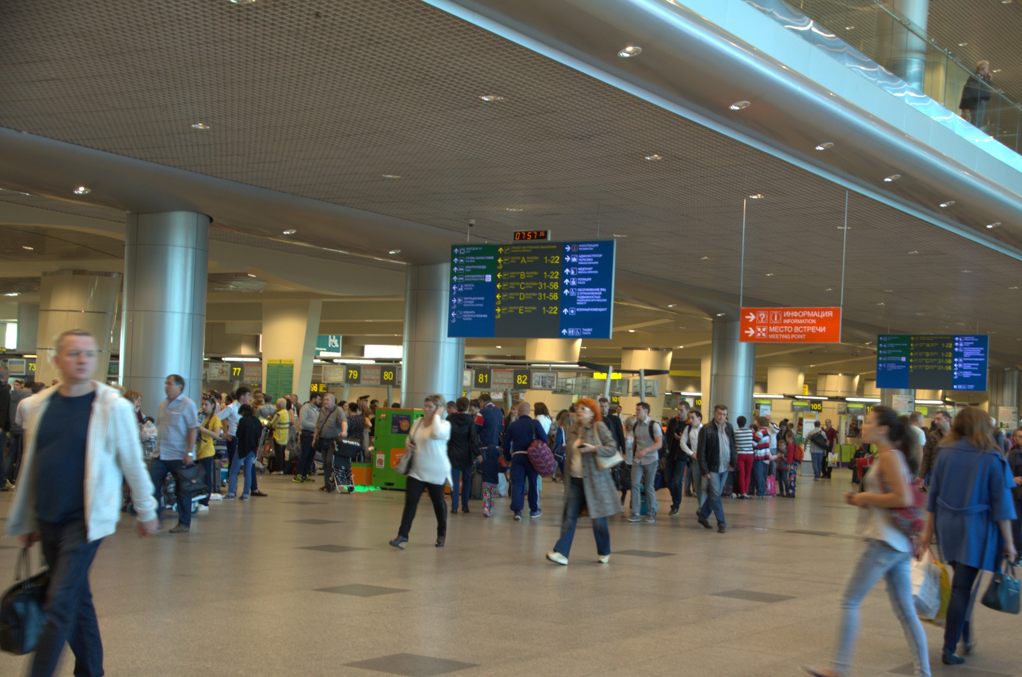 Menschen mit Gepäck durch den Flughafen gehen, übergeordnete Schilder, Säulen, Deckenleuchten und eine Glasbrüstung auf der rechten Seite.