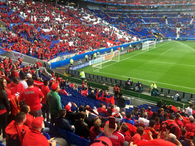 Ein volles Stadion mit Sitzplätzen und Stehplätzen, Fahnen, einem Tor und Deckenleuchten, während die Menge gespannt auf den Beginn eines Fußballspiels wartet.