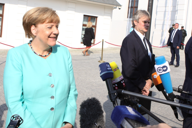 Bundeskanzlerin Angela Merkel beim Sprechen mit Reportern vor einem Gebäude mit einer Gruppe von Menschen und Equipment bei bewölktem Himmel.