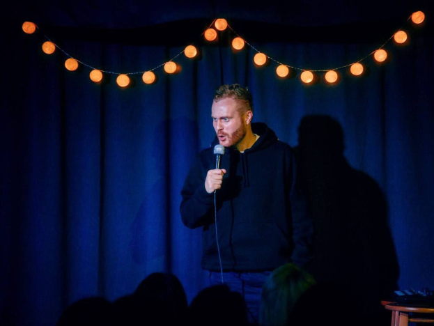 Ein Mann hält ein Mikrofon auf einer hell erleuchteten Bühne bei einem Stand-up-Comedy-Event, rechts von ihm steht ein Tisch mit Gegenständen und im Hintergrund ist das Publikum sichtbar.