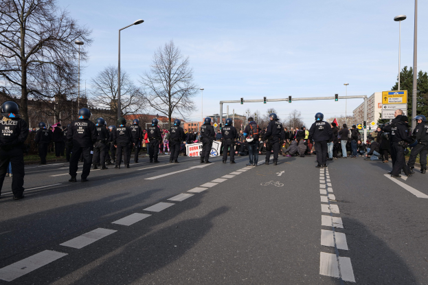 Gruppe von Polizisten in schwarzen Uniformen und Helmen, die auf der Seite einer Straße mit Laternen, Verkehrszeichen, Bäumen, Gebäuden und einem klaren blauen Himmel im Hintergrund stehen.