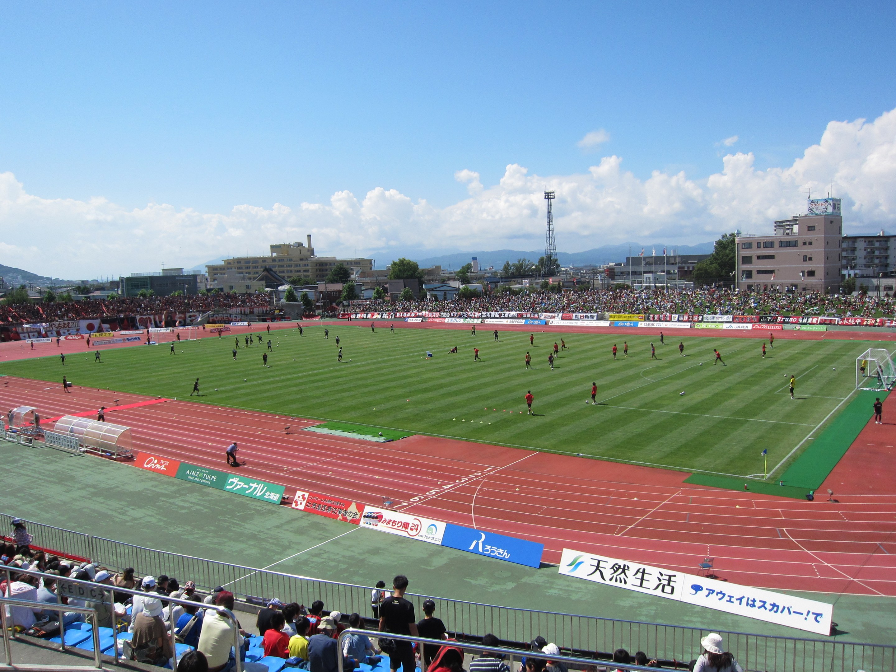 Ein großes Stadion voller Zuschauer bei einem Fußballspiel mit sitzenden und stehenden Zuschauern, Geländern, Beschriftungen und umgeben von Gebäuden, Bäumen, Türmen, Hügeln und einem bewölkten Himmel.