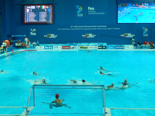 Eine Gruppe von Menschen, die Wasserball in einem großen Pool mit einem Netz in der Mitte spielen, Zuschauer sitzen auf Stühlen auf der linken Seite und eine Wand im Hintergrund zeigt Text und Logos für die 2016 FINA-Weltmeisterschaften in Kazan.
