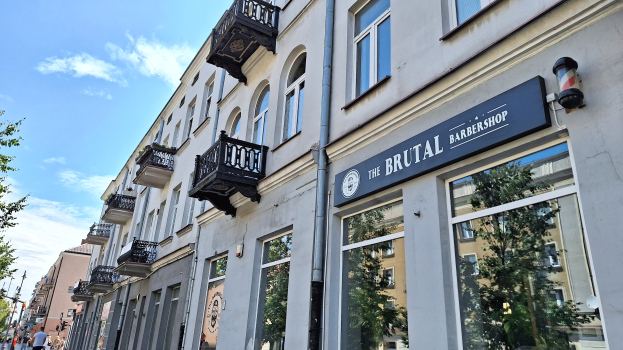 Brutalistischer Barbershop-Außenbereich in Berlin mit eckigen Betongebäuden, Fenstern, Geländern, Balkonen, Namensschildern, Bäumen, Straßenlaternen, Passanten und bewölktem Himmel.