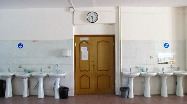 Ein Badezimmer mit mehreren Waschbecken, bodenebene Mülltonnen, einer Tür im Hintergrund, einer Wanduhr und einem Spiegel auf der rechten Seite.