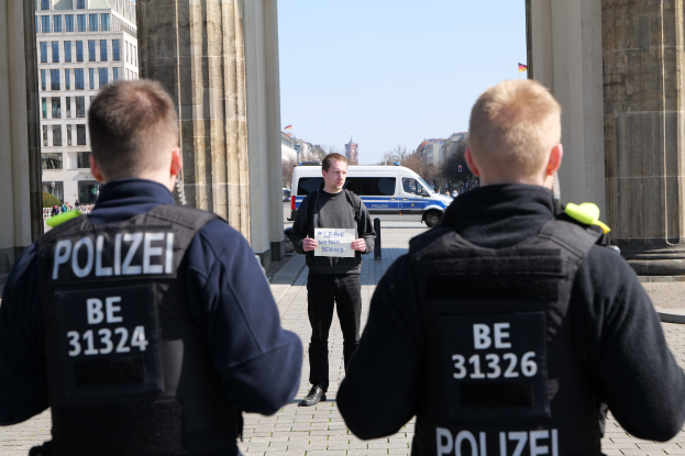 Drei Polizeibeamte stehen vor einem Gebäude, mit einem Mann, der ein Papier zwischen ihnen hält, flankiert von Säulen, während Fahrzeuge, Menschen, Bäume und Gebäude den Hintergrund unter einem klaren Himmel füllen.