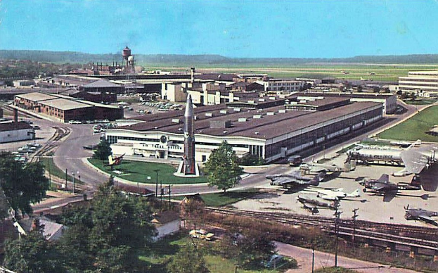Altes Schwarz-Weiß-Foto eines Flughafens mit parkenden Flugzeugen, Gebäuden, Bäumen, Straßen, Fahrzeugen, Strommasten, Gras, Bahnschienen, Türmen, Hügeln und einem klaren blauen Himmel, mit Text entlang der unteren Kante.