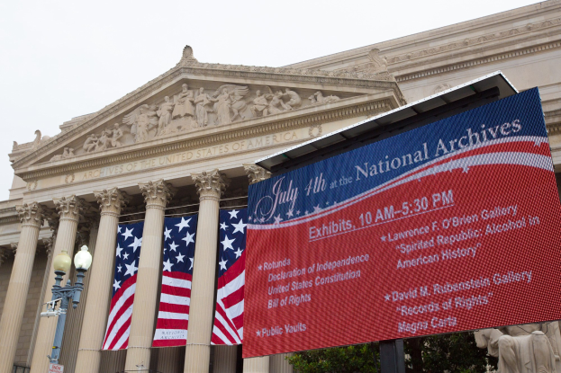 Außensicht des National Archives Gebäudes in Washington, DC, mit einer Fahne, Flaggen, Leuchten, Bäumen und Skulpturen unter einem klaren blauen Himmel.