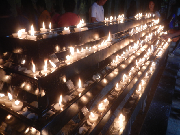 Eine Gruppe von Menschen, die um eine lange Reihe von brennenden Kerzen in einer Kirche stehen, mit einer Wand im Hintergrund.