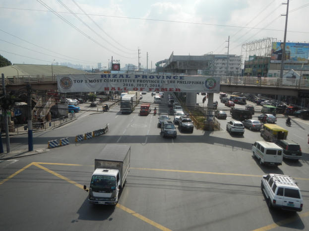 Eine belebte Straße mit zahlreichen Autos und Lastwagen, eine Brðcke im Hintergrund, Strommäste mit Drähten, eine Werbetafel mit Text, Gebäude, Bäume und ein bewölkter Himmel.