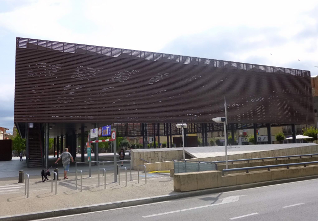 Großes Gebäude mit zahlreichen Fenstern, das als neuer Eingang zur Stadtmitte dient, umgeben von Straßeninfrastruktur, Fahrzeugen, Fußgängern, Bäumen und einem bewölkten Himmel.
