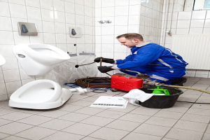 Ein Mann in einer blauen Uniform sitzt auf dem Boden vor einem Urinal und hält eine Pfeife, umgeben von Reinigungsmitteln, mit Urinalen an der linken Wand und einem Fenster auf der rechten Seite.
