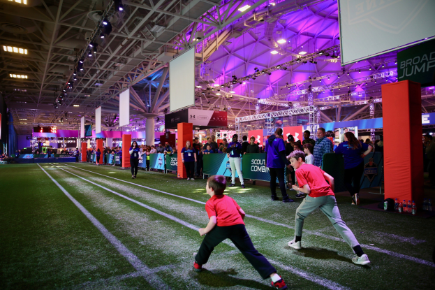 Zwei junge Jungs beim Fußballspielen auf einem Feld mit Zuschauern, umgeben von Flaschen und Gegenständen auf der rechten Seite und einem Zaun, Bannern, Anzeigebildschirm, Metallrahmen, Dach mit Lampen und Metallpfosten im Hintergrund.