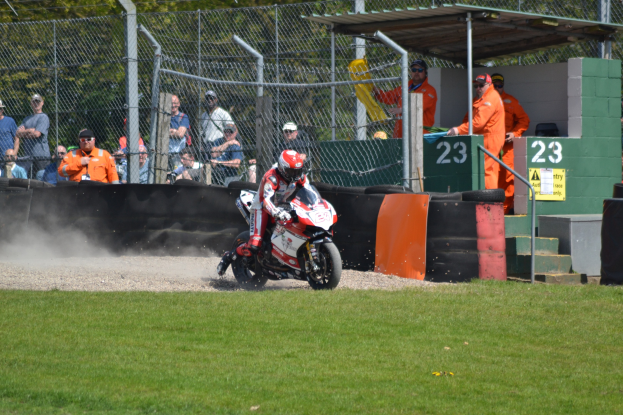 Ein Motorradfahrer mit Helm und Handschuhen fährt über ein grasbewachsenes Feld, mit Zuschauern in Mützen und Schutzbrillen, einem Zaun, Treppen mit Geländern und einem nummerierten Schuppen im Hintergrund.