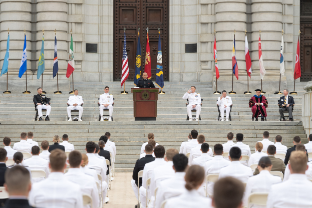 Gruppe von Menschen in weißen Uniformen auf einer Treppe vor einem Gebäude mit einem Podium, das von Fahnen flankiert ist, während einer Abschlussfeier.