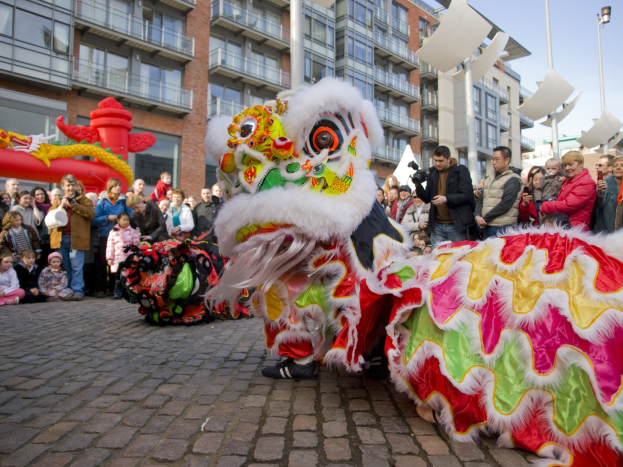 Ein farbenfrohes chinesisches Neujahrsfest in Amsterdam mit einer Löwen-Tanz-Show vor einer Zuschauermenge, darunter einige, die Fotos machen, vor einer Kulisse aus Gebäuden, Laternenmasten und einem klaren blauen Himmel.