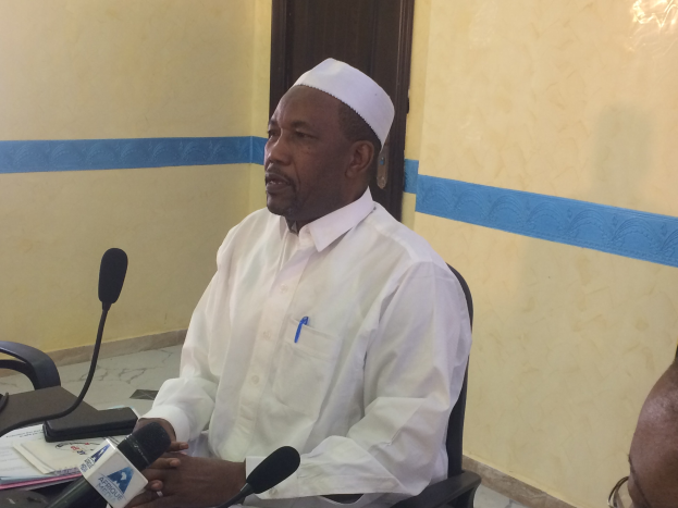 Ein Mann in weißem Hemd und Hut sitzt an einem Tisch mit Mikrofonen, spricht, während Papiere und Gegenstände auf dem Tisch zu sehen sind, mit einem anderen Mann mit Brille zu seiner Rechten, in einem Raum mit einer Wand und einer Tür im Hintergrund.