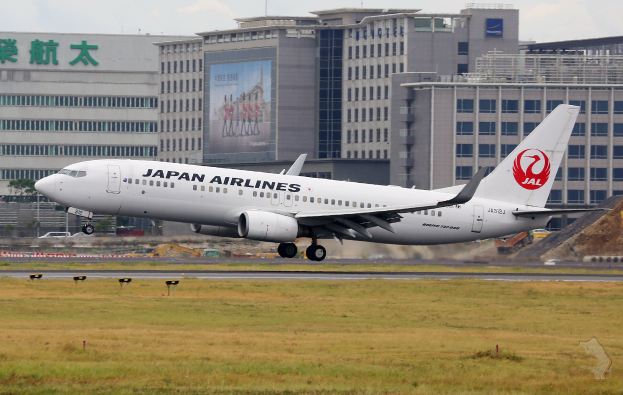 Ein Japan Airlines Airbus A320-200 beim Start von Tokyo Narita International Airport, umgeben von grünem Gras mit Gebäuden und Fahrzeugen im Hintergrund unter einem teilweise bewölkten Himmel.
